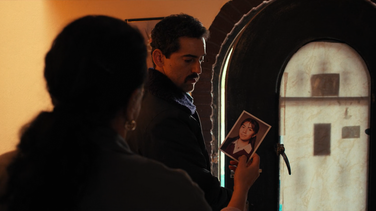 An older woman is showing a picture of a young woman to a man. Both are in a doorway and the man is already leaving.