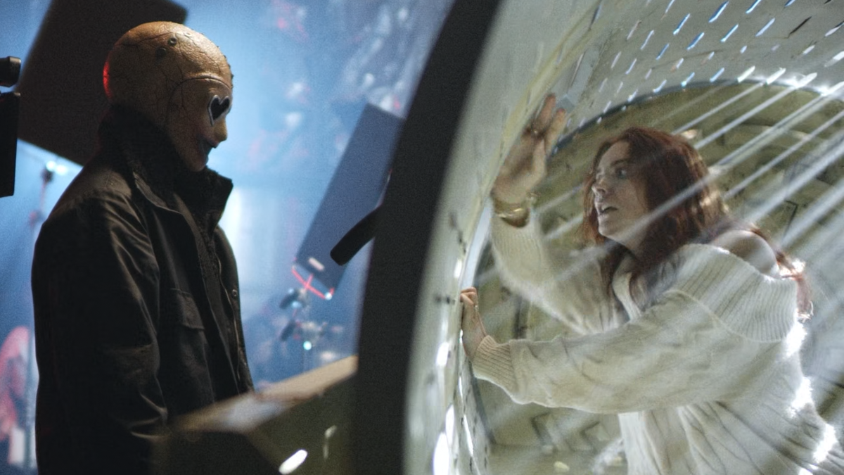 A man in all black clothes and a creepy looking skull mask is looking inside a machine where a woman is unsuccessfully trying to hide from the said man.