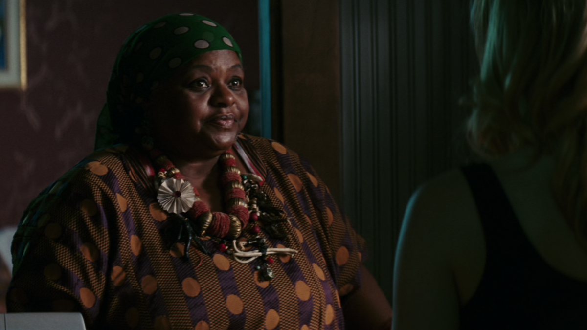 Caroline is looking at Mama Cynthia who is a Hoodoo priestess. Mama Cynthia is an older black woman. She has a strange necklace around her neck.