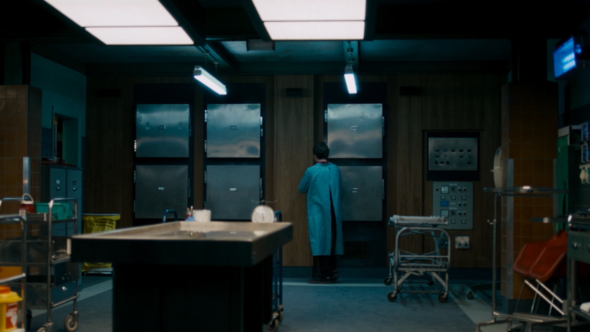 Austin Tilden is working in their morgue. He is standing near morgue drawers. There is an autopsy table in the foreground.