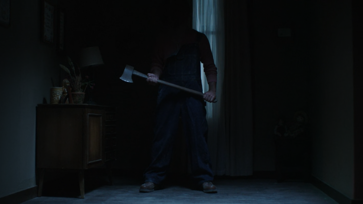 In a dark hallway a man is standing holding an axe. You can’t see his face because it’s covered by shadows.