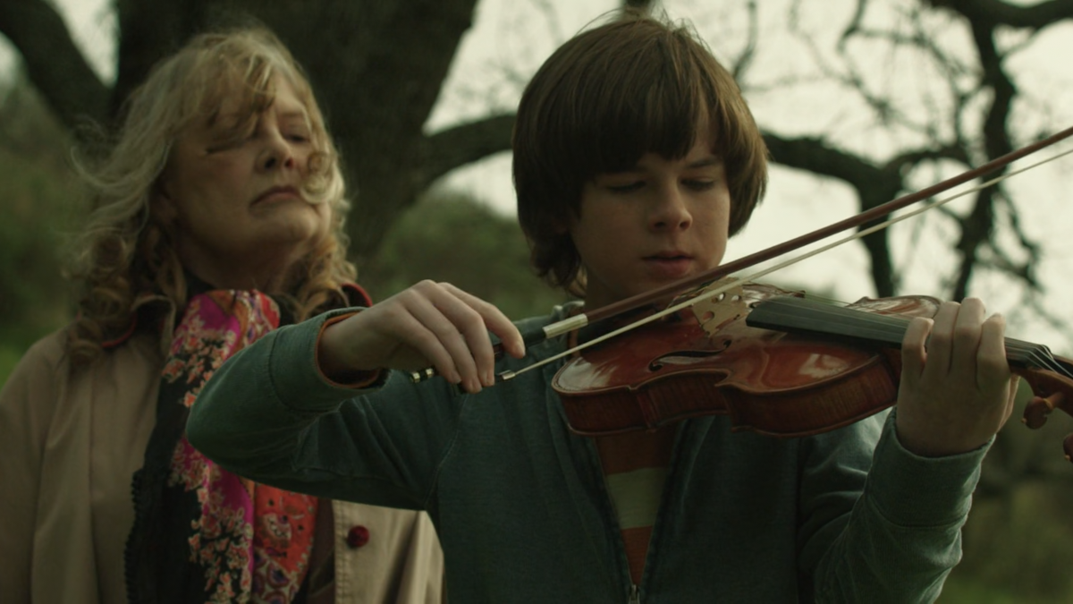 George is playing a violin. His grandmother is watching him play from the behind.