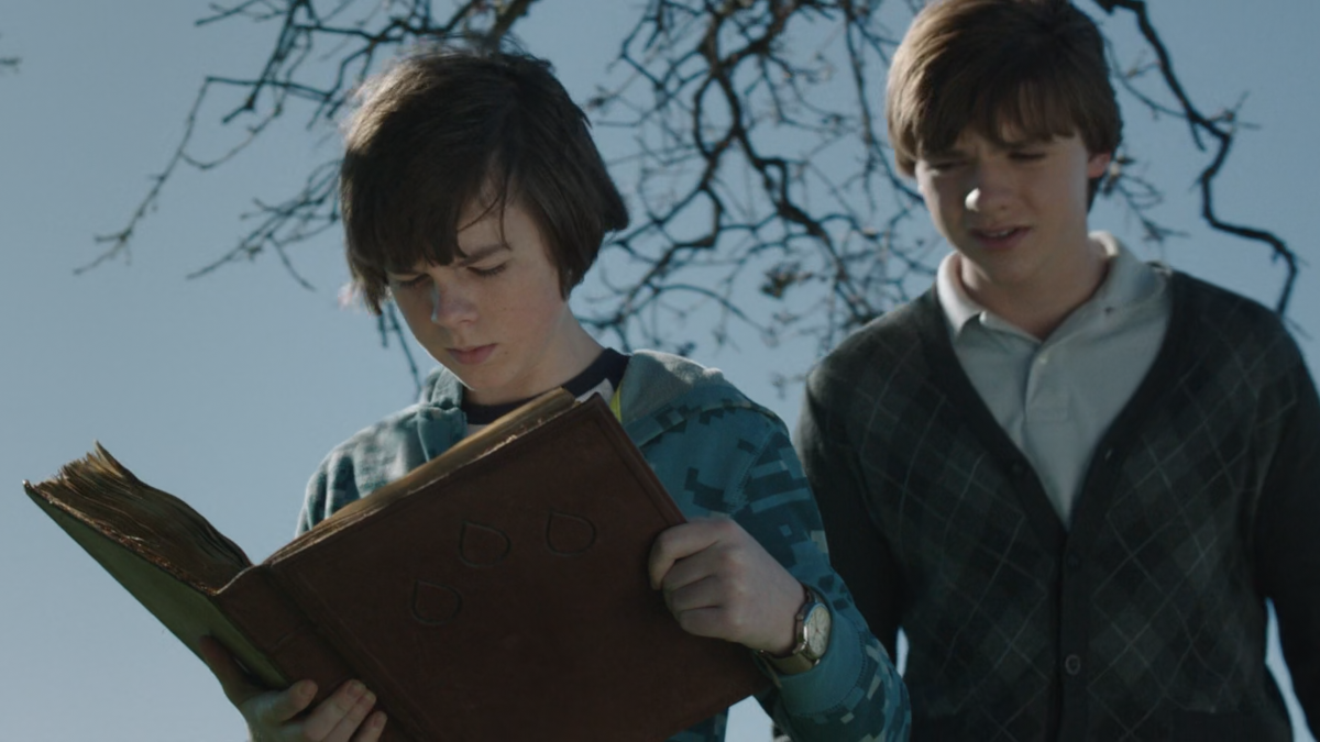 George is holding a large book. This leather-bound book looks old. There are three tear shaped marks on its front cover. George’s brother Buddy is peering over George’s shoulder to look at the book.